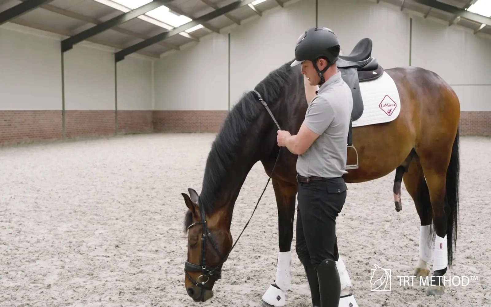 A bay horse stretching its neck down and releasing tension while Tristan Tucker stands at the saddle